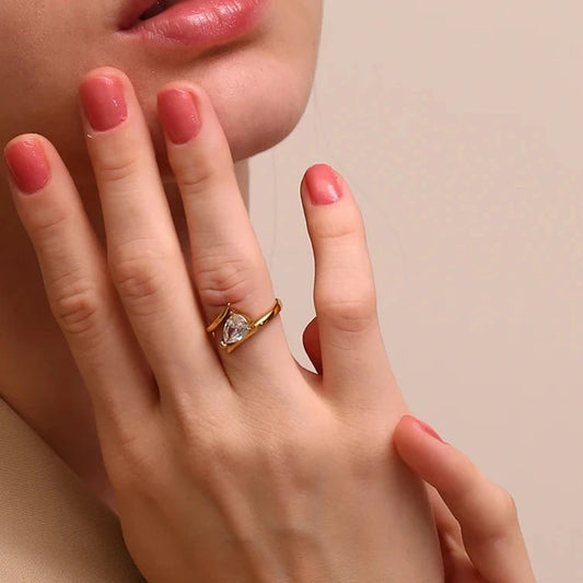 Close-up of a hand with a gold ring featuring a heart-shaped diamond, against a neutral background.